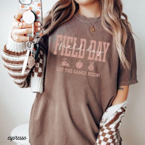 May include: Espresso-colored t-shirt with the words "Field Day" in pink script. Below the text are three small pink icons and the phrase "Let the Games Begin!" The shirt is worn with a cream-colored sweater with brown checkered sleeves.