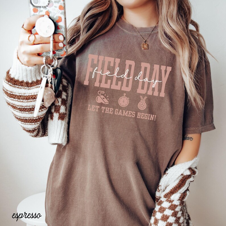 May include: Espresso-colored t-shirt with the words "Field Day" in pink script. Below the text are three small pink icons and the phrase "Let the Games Begin!" The shirt is worn with a cream-colored sweater with brown checkered sleeves.
