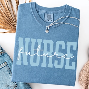 Pode incluir: Uma camiseta azul Comfort Colors com as palavras "NURSE future" impressas na frente em azul claro e branco. Um colar de prata com um pingente está drapeado sobre a camisa. A camisa está dobrada sobre uma superfície branca.