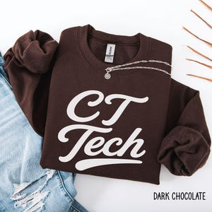 May include: A dark chocolate brown crewneck sweatshirt featuring the white text CT Tech in a cursive script font. The garment is displayed alongside light blue distressed denim jeans and a silver chain necklace with a small circular pendant.