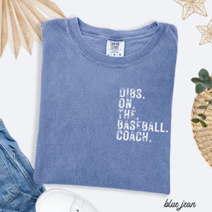 May include: A blue Comfort Colors t-shirt with the text "DIBS. ON. THE. BASEBALL. COACH." in white. The shirt is folded and has a crew neck. The words "blue jean" are written in cursive at the bottom right.