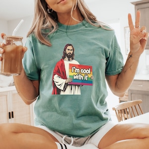 May include: A teal blue t-shirt with a graphic of Jesus holding a sign that says "I'm cool with it". The sign has a rainbow stripe across the top.