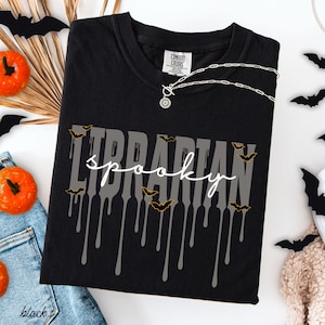 K&ouml;nnte beinhalten: Schwarzes T-Shirt mit den Worten "Librarian spooky" in Grau und Wei&szlig;, mit tropfenden Akzenten und Fledermausgrafiken. Das Shirt ist von der Marke Comfort Colors. Eine silberne Halskette mit Anh&auml;nger ist sichtbar.