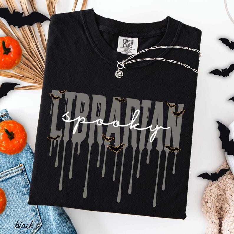 K&ouml;nnte beinhalten: Schwarzes T-Shirt mit den Worten "Librarian spooky" in Grau und Wei&szlig;, mit tropfenden Akzenten und Fledermausgrafiken. Das Shirt ist von der Marke Comfort Colors. Eine silberne Halskette mit Anh&auml;nger ist sichtbar.