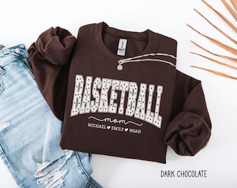Könnte beinhalten: Ein dunkelbraunes Sweatshirt mit Rundhalsausschnitt, das das Wort BASKETBALL in weißen Blockbuchstaben mit schwarzem Punktmuster zeigt, gefolgt von dem Text mom und den Namen MICHAEL, EMILY und NOAH, getrennt durch Herzen.