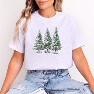 K&ouml;nnte beinhalten: Ein hellgraues T-Shirt mit einem Aquarelldesign von drei Weihnachtsb&auml;umen. Die B&auml;ume sind gr&uuml;n mit wei&szlig;em Schnee und rosa Schleifen. Das Shirt hat einen Rundhalsausschnitt und kurze &Auml;rmel.