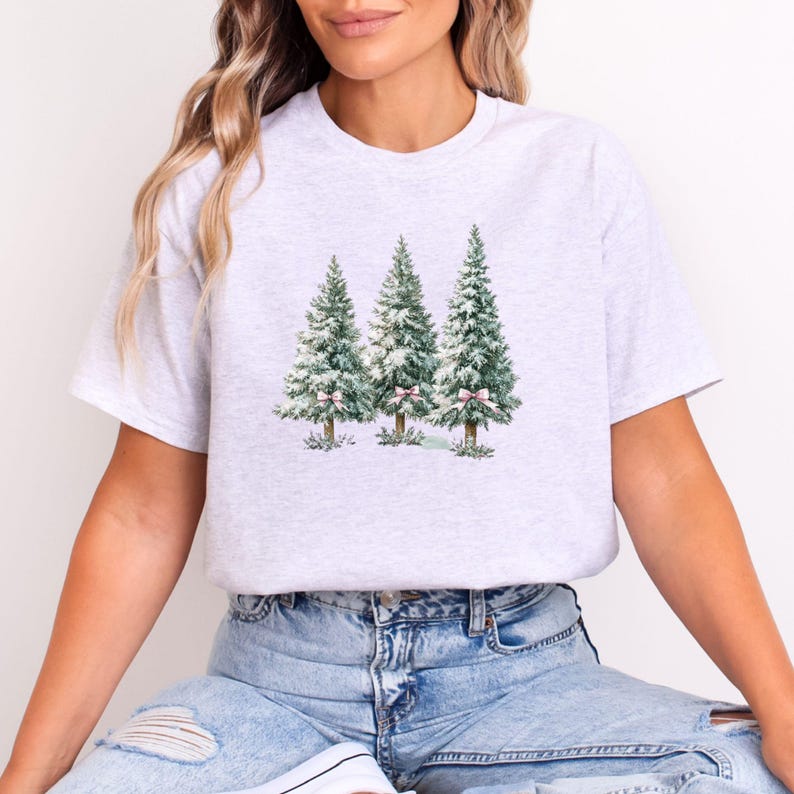 K&ouml;nnte beinhalten: Ein hellgraues T-Shirt mit einem Aquarelldesign von drei Weihnachtsb&auml;umen. Die B&auml;ume sind gr&uuml;n mit wei&szlig;em Schnee und rosa Schleifen. Das Shirt hat einen Rundhalsausschnitt und kurze &Auml;rmel.