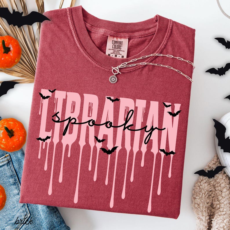 K&ouml;nnte beinhalten: Bordeauxfarbenes T-Shirt mit den Worten "LIBRARIAN spooky" in Rosa und Schwarz, mit tropfendem Farbeffekt und Fledermaus-Silhouetten. Eine silberne Halskette mit Anh&auml;nger befindet sich auf dem Shirt. Halloween-Thema.