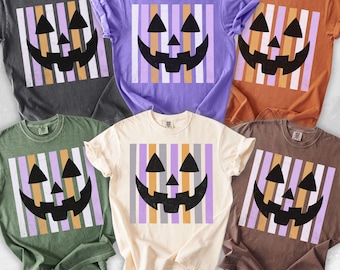 May include: A collection of five short-sleeved t-shirts in pepper, violet, yam, moss, and ivory colours. Each shirt features a central graphic of a black jack-o'-lantern face layered over a background of vertical stripes in light purple and orange.