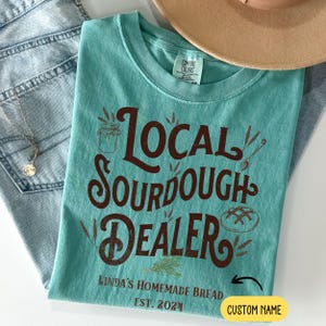 Peut inclure: Un t-shirt bleu turquoise avec un graphisme brun qui dit "Local Sourdough Dealer" avec un pot de levain, des &eacute;pis de bl&eacute; et un pain. Le texte sous le graphisme dit "Linda's Homemade Bread Est. 2024" et "Custom Name".