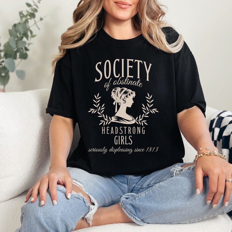 May include: A black t-shirt with a white graphic design that says "Society of obstinate Headstrong Girls seriously displeasing since 1813". The graphic features a silhouette of a woman with her hair up in a bun.