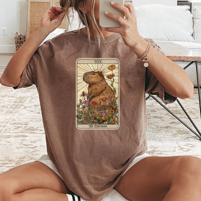 Pode incluir: Uma camiseta marrom com um gr&aacute;fico de uma carta de tar&ocirc; que apresenta uma capivara cercada de flores. A carta &eacute; rotulada como "XIX O Capibara".