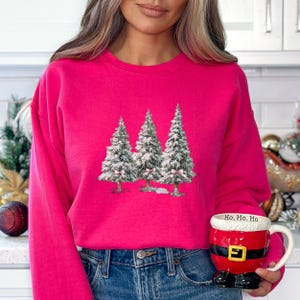 K&ouml;nnte beinhalten: Ein leuchtend pinkfarbenes Sweatshirt mit einer Grafik von drei schneebedeckten Weihnachtsb&auml;umen mit rosa Schleifen. Das Sweatshirt wird mit einer blauen Jeans kombiniert. Eine Tasse im Santa-Thema mit "Ho, Ho, Ho" wird gehalten.
