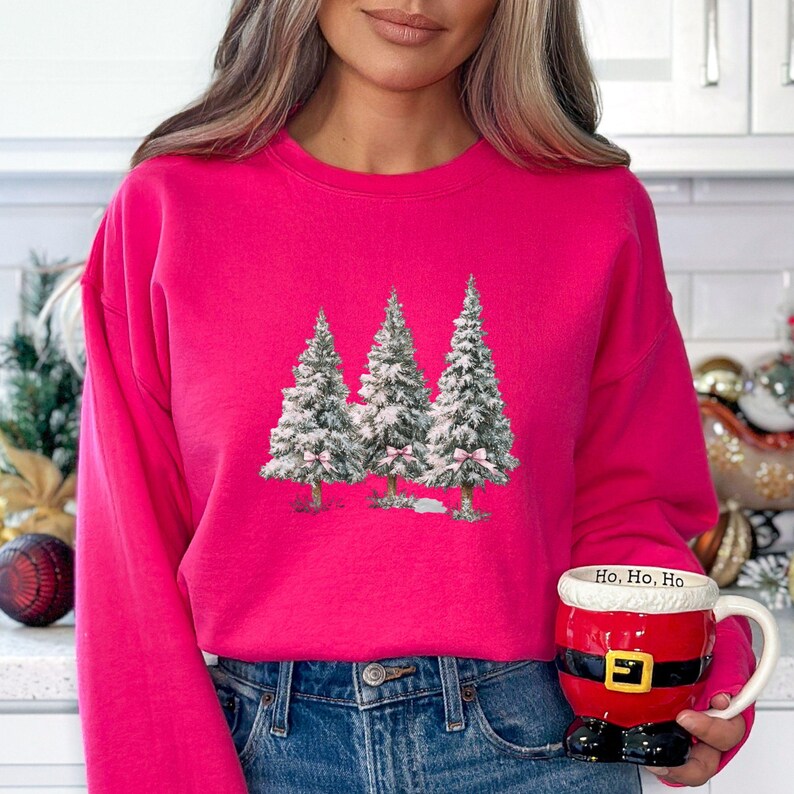 K&ouml;nnte beinhalten: Ein leuchtend pinkfarbenes Sweatshirt mit einer Grafik von drei schneebedeckten Weihnachtsb&auml;umen mit rosa Schleifen. Das Sweatshirt wird mit einer blauen Jeans kombiniert. Eine Tasse im Santa-Thema mit "Ho, Ho, Ho" wird gehalten.