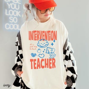 May include: Ivory-colored t-shirt with the words "INTERVENTION TEACHER" in red. The shirt features blue illustrations of a cloud, apple, heart, and a duck with a chalkboard. The text "it's a good day to teach" is also included.