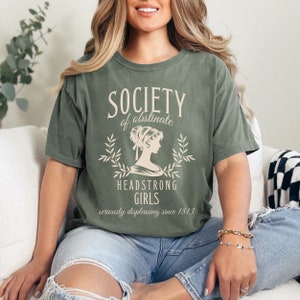 May include: A green t-shirt with the text "Society of obstinate Headstrong Girls seriously displeasing since 1813" and a vintage illustration of a woman.