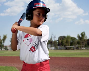 Op de afbeelding: Een jong persoon draagt een blauwe en witte baseballhelm met een rood en wit logo, een wit t-shirt met een rood en wit cartoon gezicht en een rode broek. Hij/zij houdt een houten honkbalknuppel vast en maakt zich klaar om te slaan.