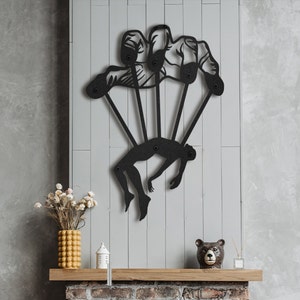 Könnte beinhalten: Schwarze Wandskulptur aus Metall, die eine Person darstellt, die von einer Hand mit fünf Fingern gehalten wird. Die Skulptur ist an einer grauen Wand mit einem gemauerten Kamin darunter montiert.