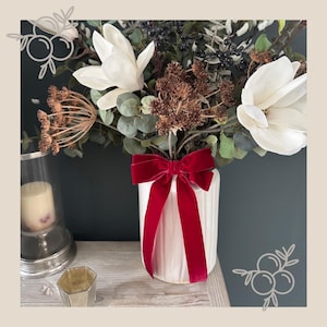 May include: A white, ribbed vase with a deep red velvet bow contains a floral arrangement. The arrangement features large white magnolia flowers, green foliage, and brown accents. The vase sits on a wooden surface against a dark green backdrop.