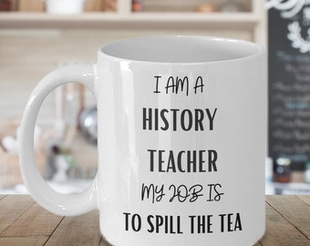 May include: White ceramic coffee mug with black text that reads "I am a history teacher my job is to spill the tea."
