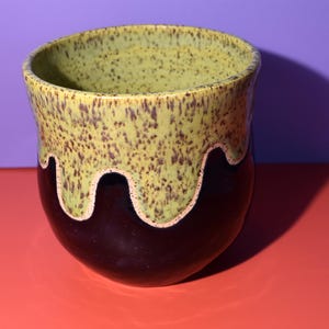 May include: A ceramic mug featuring a two-tone glaze design. The upper portion is a speckled light yellow-green, while the lower portion is a solid dark brown. The glaze application creates a distinct, wavy drip pattern where the two colors meet.