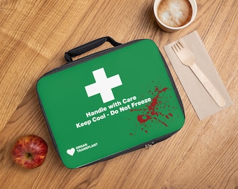 May include: A rectangular green lunch bag with a black handle and zipper. The front features a white cross, red blood splatter graphics, and the text: Handle with Care, Keep Cool - Do Not Freeze, and Organ Transplant with a heart icon.