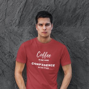 以下が含まれることがあります： 白い文字で「Coffee in one hand Confidence in the other」と書かれた赤いTシャツです。