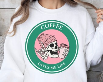 May include: White sweatshirt with a circular graphic featuring a skeleton wearing a pink beanie and holding a coffee cup. The graphic is surrounded by a green border with the words "COFFEE GIVES ME LIFE".