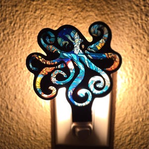May include: A decorative night light featuring a colorful octopus design. The octopus is black with blue, orange, and yellow accents. The night light is plugged into a wall outlet and emits a soft glow.
