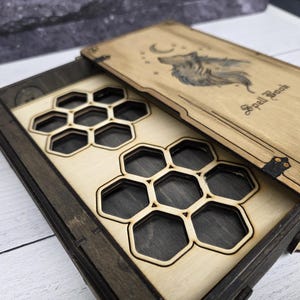May include: Wooden box with a sliding tray featuring a honeycomb design. The lid is engraved with a wolf and crescent moon, with the words "Spell Book". The box is made of light and dark wood.