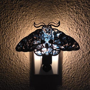 May include: A decorative night light shaped like a moth, featuring a black silhouette with intricate wing patterns. The wings have a blue and white stained glass effect, and the light plugs directly into a wall outlet.
