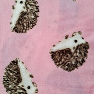 May include: Pink fabric with a repeating pattern of hedgehogs. The hedgehogs are white and brown with black eyes and noses. The fabric has a soft, fuzzy texture.