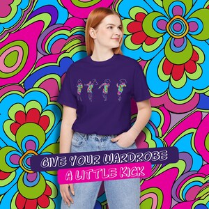 Peut inclure: Un t-shirt violet avec un motif de quatre femmes dansant dans un style rétro. Le texte "Give your wardrobe a little kick" est imprimé sur une bannière rose sous le motif.