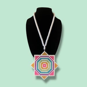 Puede incluir: Un collar de cuentas de colores con un colgante cuadrado grande. El colgante presenta un diseño geométrico con cuentas rosas, naranjas, amarillas, verdes y azules. El collar se exhibe en un maniquí negro.