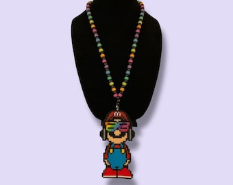 May include: A pixelated Mario pendant necklace with a rainbow beaded chain. The pendant features Mario wearing headphones and rainbow-colored glasses, with red overalls, a red hat, and red shoes. The necklace is displayed on a black mannequin.