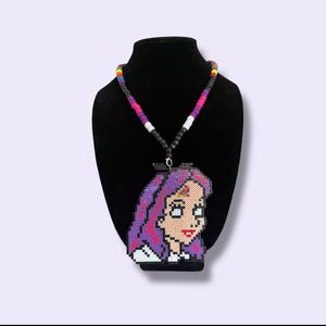May include: A beaded necklace featuring a pendant with a cartoon portrait of a woman with purple hair. The pendant is made of colorful beads. The necklace has a black cord with colorful beads.