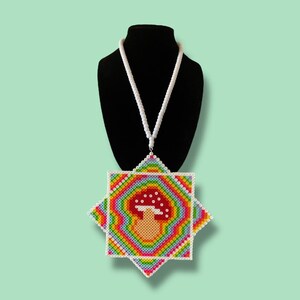 Puede incluir: Un collar de cuentas colorido con un diseño de hongo. El colgante es cuadrado con un contorno en forma de estrella, mostrando un degradado arcoíris. El hongo tiene un sombrero rojo con puntos blancos. El collar se exhibe en un maniquí negro.