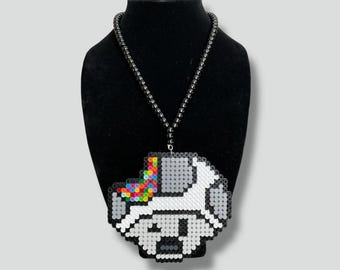 May include: A pixelated pendant necklace featuring a white unicorn with a rainbow mane and tail. The pendant is made of small, colorful beads. The necklace has a black beaded chain and is displayed on a black mannequin.