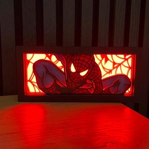 Può includere: Un decoro da parete luminoso in legno rosso e nero con una silhouette di Spiderman in una ragnatela. La luce brilla attraverso il design ritagliato, creando un bagliore rosso.