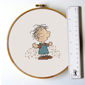 May include: Cross-stitch embroidery of a cartoon character in a wooden hoop. The character has messy blonde hair, a blue outfit, and brown shoes. The embroidery is on a white fabric. A ruler is next to the hoop, measuring up to 6 inches.