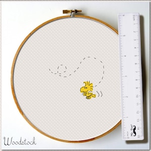 May include: A white embroidery hoop with a yellow and black embroidered image of the Peanuts character Woodstock. The embroidery is of Woodstock flying with a dotted line trail behind him. The hoop is 15 cm in diameter and is sitting on a white surface next to a ruler.