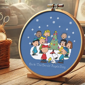 May include: A blue and white cross-stitch embroidery hoop featuring a scene of the Peanuts characters singing carols around a Christmas tree. The text "Hark! The Herald Angels Sing" is at the bottom of the hoop.