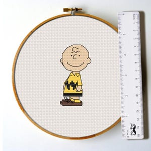 May include: A cross-stitch pattern of Charlie Brown from Peanuts wearing his signature yellow shirt with black zigzags. The pattern is framed in a wooden embroidery hoop.