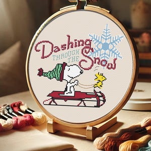 Könnte beinhalten: Ein runder Holzstickrahmen mit weißem Stoffhintergrund. Der Stoff zeigt ein Kreuzstich-Design von Snoopy in einem roten Schlitten mit einem gelben Vogel. Der Text "Dashing Through The Snow" befindet sich über Snoopy.
