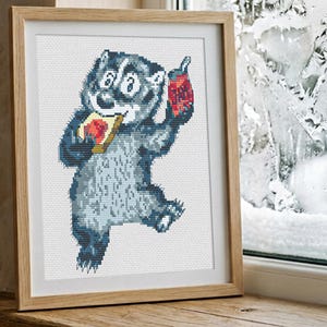 May include: Cross-stitch art of a cartoon badger holding a piece of toast with jam and a jar of jam. The badger is in shades of blue and gray, framed in a light wood frame. The background shows a window with a snowy scene.