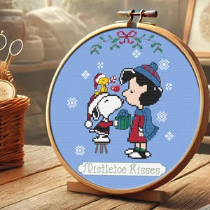 May include: A blue and white cross-stitch embroidery hoop with a scene of Snoopy and Lucy from Peanuts. Snoopy is wearing a red Santa hat and holding a gift. Lucy is wearing a red dress and a blue scarf. The text "Mistletoe Kisses" is written below the characters.