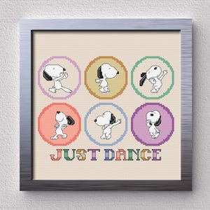 May include: Cross-stitch artwork featuring Snoopy in various dance poses, each within a colorful circle. The phrase "JUST DANCE" is stitched in rainbow colors below the circles. The artwork is framed in a silver-colored frame.