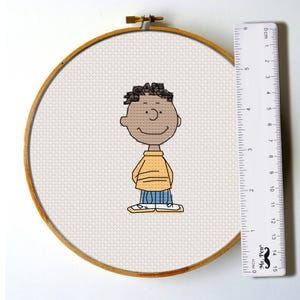 May include: Cross stitch pattern of a cartoon character with brown skin, black hair, a yellow shirt, blue pants, and white shoes. The pattern is displayed in a wooden embroidery hoop.