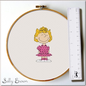 May include: A cross-stitch pattern of Sally Brown from the Peanuts comic strip. The pattern shows Sally in a pink polka dot dress with her signature blonde hair.