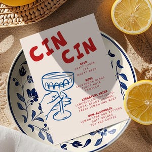 以下が含まれることがあります： 赤色の「CIN CIN」という文字が入ったメニューカード。ビール、ワイン、シグネチャードリンク、ノンアルコール飲料が記載されています。カードは青と白の花柄のプレートの上にあり、レモンの半分と丸ごとのレモンが近くにあります。